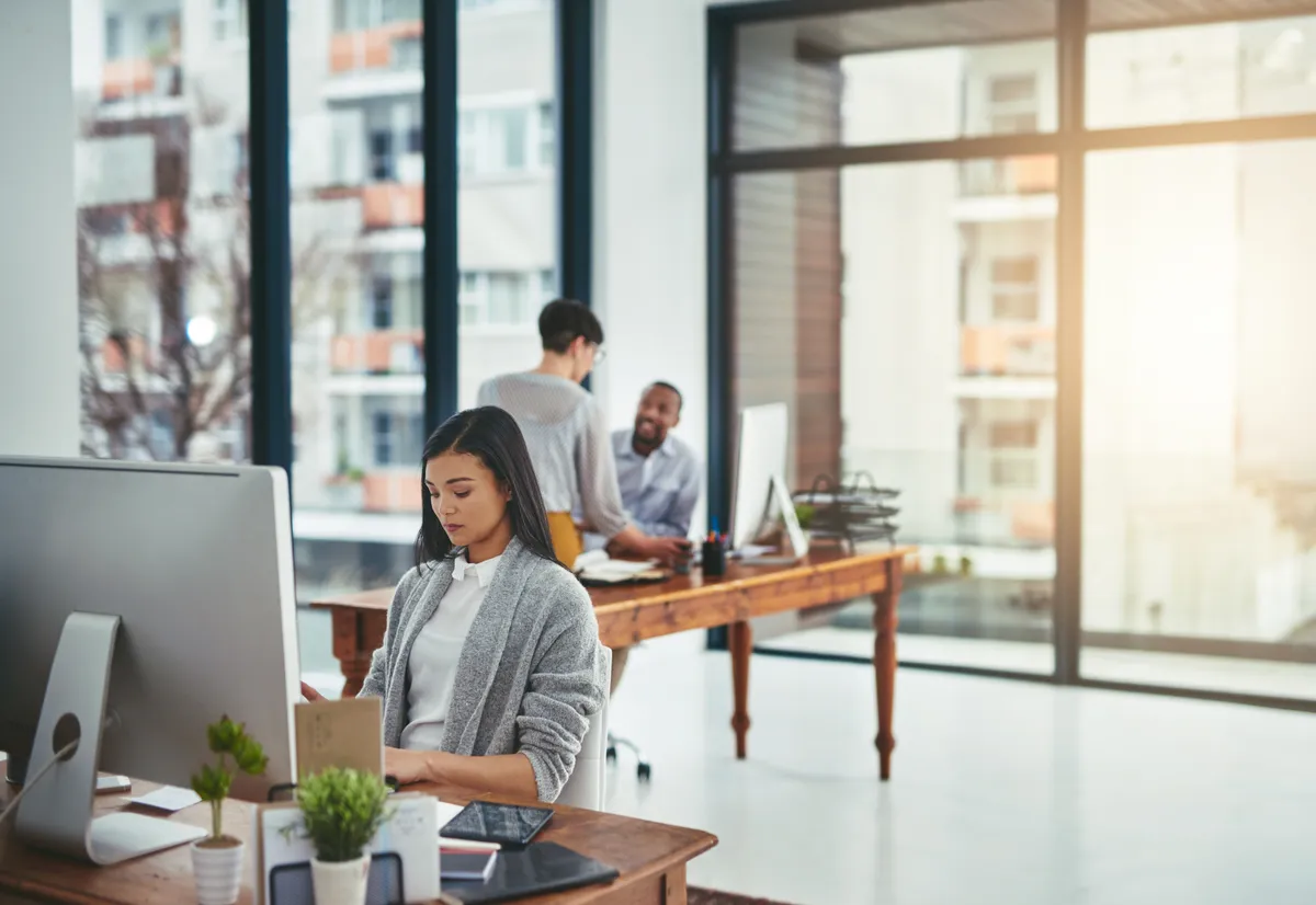 Bright open-plan office where a woman works at a desktop computer while two colleagues talk behind her